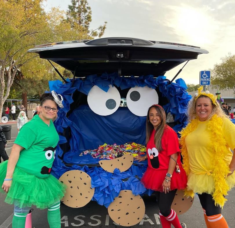 Volunteers in colorful costumes beside a decorated car at Trunk or Treat